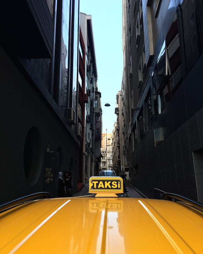 A yellow taxi navigates a narrow urban alley with tall buildings on either side, showcasing city life.