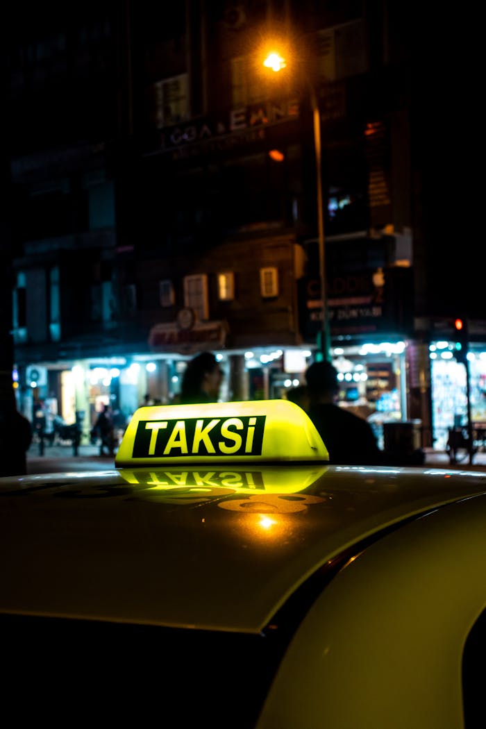 Yellow taxi sign reading 'TAKSI' glowing at night with cityscape background.